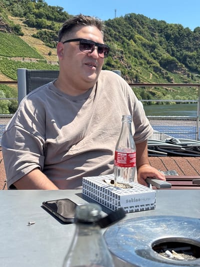 A man wearing sunglasses and a beige t-shirt sits at an outdoor table with a bottle of Coca-Cola, a box of tissues, and a smartphone. In the background, a river flows past a hillside covered in vineyards. This scene is set in Piesport, Germany, a town renowned for its wine production. The man appears relaxed, enjoying the sunny day and the scenic view. The presence of an ashtray suggests a casual, possibly smoking-friendly environment.