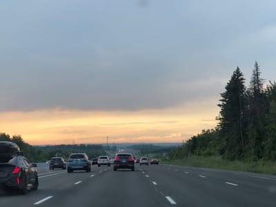 This media file shows a view from a car driving on a highway in King, Canada. The scene depicts a multi-lane highway with several cars ahead, including a black SUV with a roof carrier, a blue SUV, a black SUV, and a white SUV. The highway is bordered by trees and greenery on the right side, and a rolling landscape with more trees and some distant structures on the left. The sky is overcast with soft clouds, and the light suggests it is either early morning or late evening, with a warm glow on the horizon indicating sunrise or sunset. The overall mood is peaceful and serene, typical of a highway drive through a scenic rural area. There is no visible text in the image.