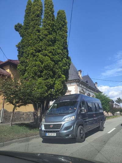 A dark blue camper van with the license plate HP IK 19 is traveling on a paved road in Sighetu Marmatiei, Romania. The van is on the right side of the road, indicated by the white line marking the lane. The van has the brand name "ADRIA" visible on its side and front. On the left side of the road, there is a large, mature evergreen tree with a distinctive shape, resembling a hand with outstretched fingers. Behind the tree, a house with a yellow facade and a brown tiled roof is visible. Further in the background, another building with a dark, pitched roof and architectural details is present. The sky is clear and blue with a few scattered white clouds. The scene appears to be during daylight hours.