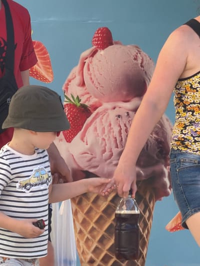 The image depicts two people interacting with a large, flat depiction of ice cream and waffle cone. In the foreground, a child wearing a grey cap and a striped shirt is lying down, looking towards the right. The child is holding a small dark object in their right hand. Behind the child, an adult's arm is visible, reaching towards a bottle of dark liquid on the waffle cone. The adult is wearing a floral print top and denim shorts. The background is a plain blue surface, suggesting an indoor setting or a backdrop. The lighting appears to be even, with no strong shadows, indicating it's likely daytime or under artificial lighting. The scene evokes a playful or relaxed atmosphere, possibly related to a promotional event or an artistic display. No visible text is present in the image.