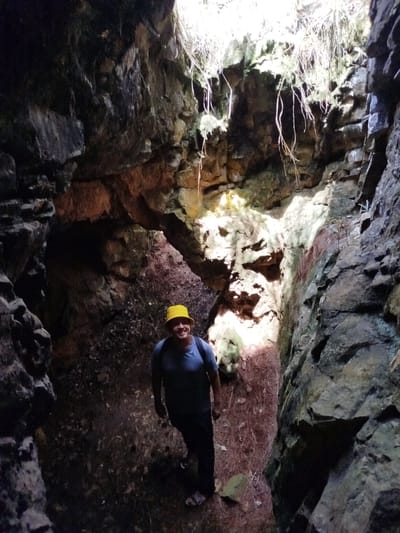 The image captures a male individual standing within a narrow, natural rock crevice or cave entrance, stated to be in Đa Thọ, Vietnam. The man, appearing middle-aged, is positioned centrally, facing the camera with a distinct smile. He is wearing a bright yellow bucket hat, a blue short-sleeved t-shirt, dark pants, and carries a dark backpack. The geological formation enclosing him consists of steep, rugged rock walls, predominantly dark grey and brown, with visible stratification and fractured surfaces. Sections of the rock display reddish-brown hues and lighter green patches, indicative of mineral deposits or moss. Natural light filters intensely from a narrow opening directly above the crevice, brightly illuminating the upper rock faces and sparse, pale vegetation, including hanging roots and dried grasses, that descend from the opening. The ground on which the man stands is covered with a layer of reddish-brown pine needles, dry leaves, and small scattered rocks. The overhead light source creates deep shadows on the lower parts of the crevice and around the individual, highlighting the rough textures of the rock walls.