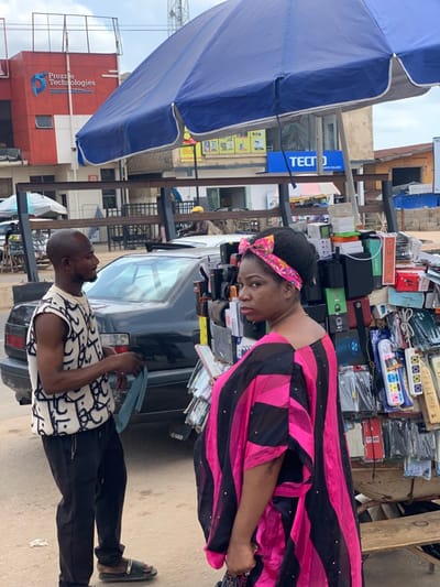 The scene is a street market in Ondo, Nigeria, located at coordinates latitude: 7.252, longitude: 5.199. A woman in a pink and black dress with a pink headband is standing behind a mobile stall, while a man in a white and black patterned sleeveless shirt and black pants stands to the left of the stall. A black car is parked behind the stall. The woman is looking towards the left of the frame, while the man is looking to his right. The stall is full of various electronics, including cell phone cases, chargers, and power adapters. A blue umbrella is above the stall, providing shade for the vendor and customers. The background of the image includes a building with a sign for "Prezzie Technologies". Several people are visible in the background, but their details are blurred and difficult to make out. The scene appears to be in broad daylight.