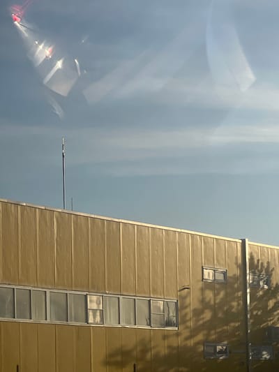 The image shows the side of a large, yellow-paneled building with a row of windows running horizontally across its facade. A tall, thin antenna stands against a clear blue sky, which is partially obscured by reflections and light streaks. Shadows of trees are cast across the building, indicating it is daytime and sunny. The building appears to be located in an industrial or commercial area, with no immediate signs of residential activity or prominent landmarks. The town of Dragushinovo, Bulgaria, is mentioned as the location, but no specific features within the image confirm this.