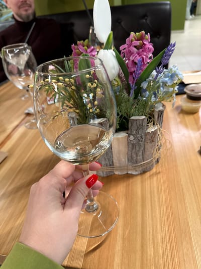 A close-up shot depicts a hand with red fingernails holding a wine glass filled with flowers. The floral arrangement consists of purple hyacinths, blue flowers, a white tulip, and other greenery. The flowers are set in a decorative wooden container bound with twine. Two other wine glasses are visible, one near the bottom of the frame and another with a white object next to it in the background. The table has a wood grain pattern. A person is partially visible in the lower left corner. The image was taken in Peresvet, Russia.