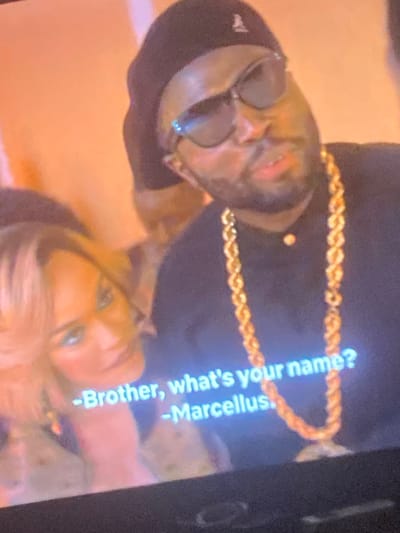 An image captures a rotated scene from a screen, displaying two individuals and subtitle text. The image is rotated approximately 90 degrees clockwise. In the upper part of the frame, a man with dark skin is visible, wearing dark sunglasses, a dark baseball cap, and a thick, gold-colored chain around his neck. He has facial hair. His gaze is directed downwards and to the left (relative to the original image orientation).
Below him, partially obscured, is a woman with blonde hair, looking upwards. Her features are less distinct due to blurriness, but she appears to be light-skinned. The background is softly blurred with warm, orange-brown tones, suggesting an indoor setting.
Subtitle text is prominently displayed on the screen, indicating a dialogue:
"-Brother, what's your name?"
"-Marcellus."
The image appears to be a photograph of a digital display, evidenced by the visible text and the overall quality, suggesting it's a still from a movie, TV show, or video.