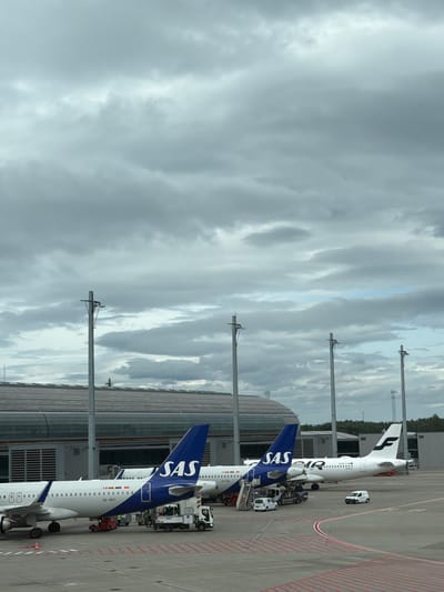 This media file shows an aerial view of airplanes parked at an airport gate. The main subjects are two SAS airplanes, identifiable by their blue and white livery and the prominent "SAS" logo on their tails. One plane is partially visible, with the registration "LN-SAE" on its tail. The other, closer to the foreground, has the registration "SE-ROY" and clearly displays "SAS". Both planes appear to be boarded by passengers, as indicated by the presence of stairs and service vehicles around them, suggesting an ongoing boarding or deplaning process.

In the background, another airplane with "FINNAIR" branding is visible. Various airport service vehicles, including baggage loaders and a small white vehicle, are positioned near the planes. The setting is an airport tarmac, with the planes parked next to a modern airport terminal building, characterized by its glass facade and horizontal wooden accents. Utility poles with visible wires stretch across the scene.

The sky is overcast with a mix of gray and lighter clouds, suggesting a possibly cloudy or overcast day, typical of Scandinavian weather. The lighting appears diffused, indicating it is daytime, though the exact time is not discernible. The overall scene depicts a routine airport operation with aircraft ready for or having completed a flight. The visible text includes airline names like "SAS" and "FINNAIR", and aircraft registration numbers "LN-SAE", "SE-ROY", and "OH-LAN". The location is identified as Gardermoen, Norway.