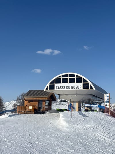 The image displays the "Casse du Boeuf" ski lift station located in a snow-covered mountain environment within the ski resort area of La Salle-les-Alpes, France. The central subject is a large, modern ski lift terminal featuring a prominent white, arched roof structure with the name "CASSE DU BOEUF" displayed in dark capital letters. Inside the station, multiple ski lift chairs, distinguished by green and black seats, are visible. The loading area in front of the station is demarcated by blue barriers and red flags. To the left and slightly behind the main lift station, a smaller, rustic wooden building with a dark, pitched roof and an attached wooden deck is present. The foreground and midground are covered in groomed snow, showing visible ski tracks. In the background, under a clear blue sky with sparse white clouds, snow-capped mountains and scattered leafless trees are visible. No people are observed in the scene.
