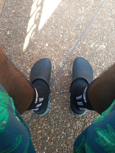 The image presents a high-angle, downward view of a person's lower legs and feet, positioned on a textured ground surface in Konongo, Ghana. The subject wears dark-toned perforated clogs, each featuring a camouflage-patterned sole, over black ankle-length socks adorned with white horizontal stripe markings. The visible portions of the legs display a dark skin tone, and the individual is wearing patterned shorts or swimwear, primarily blue with green leafy or floral designs. The ground is composed of a light brown and grey aggregate, possibly a pebbled concrete or asphalt surface. A thin, dark, linear object, resembling a cable or a seam, runs diagonally across the ground between the feet. Distinct shadows are cast on the upper left portion of the ground, indicating direct sunlight from an elevated source. No other individuals or significant objects are visible within the frame.
