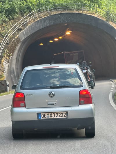 A silver Volkswagen Lupo is driving through a tunnel in Hornberg, Germany. The car is in the foreground, and the tunnel entrance is visible in the background. There is a truck with the word "KERN" on the side visible in the tunnel, and a motorcycle is behind the truck. The tunnel is lit by orange lights. The road is paved, and there are white lines on the road indicating lanes. There are trees and foliage on the embankment outside the tunnel. The license plate of the Volkswagen Lupo reads "OG XJ 2222".
