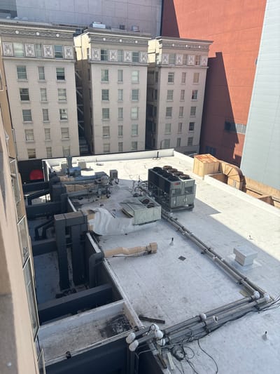 The image is a high-angle shot of a rooftop in a city, likely San Francisco, United States of America, featuring multiple buildings and extensive HVAC equipment. In the foreground, a beige-colored building facade with windows extends from the left edge. Beyond this, a flat white rooftop is visible, densely populated with various mechanical components. Prominently placed on the rooftop is a large grey HVAC unit with multiple circular fans on top. Surrounding this unit are numerous pipes, ventilation ducts, and smaller air conditioning units. Several grey metal ducts snake across the rooftop, connecting to different parts of the machinery. In the background, several taller buildings rise, some appearing to be older architectural styles with multiple floors and windows, while others are modern. One of these buildings is a reddish-brown structure with a large shadow cast across it. The sky is clear and bright, indicating daytime.