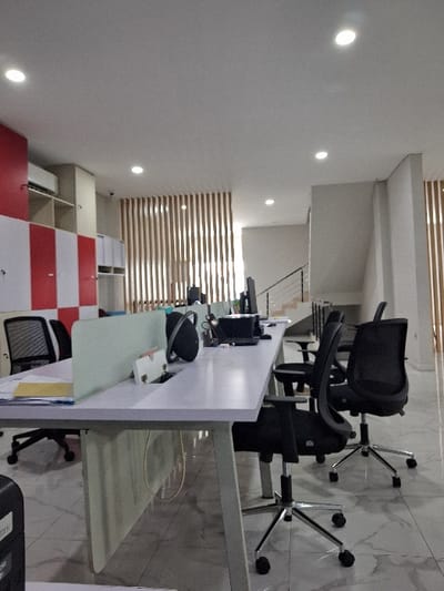 An office interior features workstations with white desks and black office chairs. A modern design incorporates a wall with horizontal wood slats, complemented by recessed lighting. The space also includes storage cabinets with white and red panels. The flooring appears to be marble.