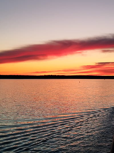 The image shows a body of water, likely a lake or wide river, at sunset or sunrise. A dark treeline silhouettes the horizon between the water and the colorful sky. The sky transitions from a pale blue/gray at the top to orange and then red near the horizon, with prominent red clouds. The water reflects the colors of the sky, creating an orange/red hue across the surface. Wave patterns are visible on the water, particularly closer to the camera, suggesting the presence of wind or movement. There is evidence of disturbances or ripples in the water at the bottom right of the frame, suggesting the movement of a boat or similar object. The image is not explicitly identified as being in Topornya, Russia, and no people or objects are identifiable in the scene.