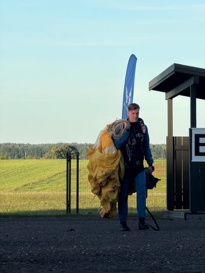 A man in a blue and black skydiver's jumpsuit, carrying a yellow parachute and its ropes, walks away from the camera on a gravel path. He is in front of a modern, dark-roofed structure with vertical wooden panels. A tall, blue flag with a white logo stands to his left. In the background, a green field stretches towards a line of trees under a clear, light blue sky. A black metal fence is visible in front of the field. The scene appears to be an outdoor drop zone, possibly at the Limbaži airfield in Limbaži, Latvia, given the context of skydivers and open fields.