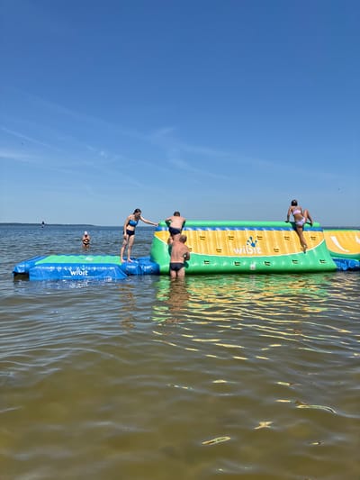 In Rewa, Poland, on a clear, sunny day, several people are engaged in activities on an inflatable water obstacle course situated in the Baltic Sea. The structure, a Wibit brand inflatable, features various sections, including a blue and green climbing wall and a yellow and green obstacle. A woman in a blue bikini is standing on the blue section, reaching towards the yellow section. Another person, wearing black swim trunks, is partially submerged in the water and appears to be climbing the yellow obstacle. A third person, also in black swim trunks, is visible from the waist up, also on the yellow section. A fourth person, a woman in a white and purple bikini, is on the top of the yellow section, with one leg extended as she climbs. In the background, a person in a white hat is standing in the water. Further in the distance, a sailboat is visible on the horizon. The water is calm with ripples and reflects the blue sky and the colors of the inflatable structure. The overall scene depicts a recreational activity at the beach.