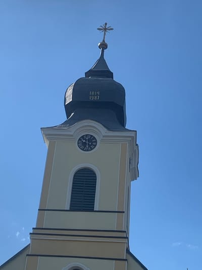 The image presents a detailed, low-angle view of a church tower in Yasinia, Ukraine, observed against a clear, cloudless blue sky. The tower's main structure is painted a light cream or pale yellow, accented with white and dark horizontal trim lines. A circular clock face with Roman numerals is visible on one side, with its hands indicating approximately 11:20. Directly below the clock, an arched window opening is fitted with dark horizontal louvers. Above this section, the tower transitions to a dark, bell-shaped, or onion-domed cupola. Inscribed on this dark cupola are two golden numerical dates: "1814" above "1987." The apex of the cupola is surmounted by an intricate golden cross, likely indicative of a Christian denomination, featuring a central sphere and radiating elements. No individuals or other structures are present in the frame.