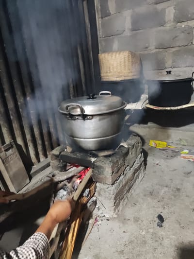 The image depicts a rustic outdoor kitchen scene, likely in Ngentrong, Indonesia, characterized by a traditional wood-fired stove. A large, metallic steamer pot, filled with food and emitting steam, is actively cooking over a fire. Adjacent to it, two dark, lidded pots are also present, one with a woven basket lid. The stove itself is constructed from bricks, with visible glowing embers beneath the steamer. A person's arm and hand, wearing a checkered sleeve, are visible on the left side of the frame, tending to the fire with sticks. The environment appears to be a simple, possibly unpaved area, with a corrugated metal wall in the background and scattered debris on the ground, including packets and what appears to be a leaf. The overall atmosphere suggests a traditional and practical approach to cooking.