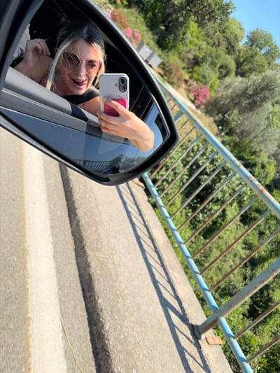 A young woman with blonde hair and sunglasses is leaning out of a car window and taking a selfie with her phone. She is sticking her tongue out at the camera. The car is driving along a road next to a blue railing. In the background, there are trees and a hillside. The road appears to be made of concrete and asphalt.