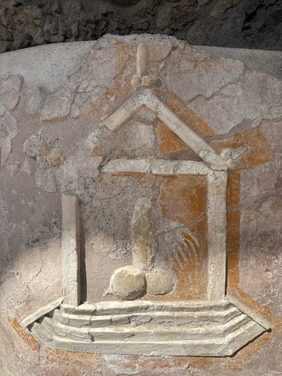 The image displays a weathered ancient fresco or shallow relief carving on a wall in Pompei, Italy. The central subject is an erect phallus with distinct testicles, framed by an aedicula, a small shrine-like architectural structure.

The aedicula is composed of a tiered, stepped base, two vertical pilaster-like supports, and a triangular pediment. Above the pediment, a smaller, upright phallic element with two spherical objects at its base is visible. Two additional spherical objects are positioned on the pediment itself, one on each side. To the right of the central phallus, a stylized hand with splayed fingers extends from the background, pointing towards the phallus.

The artwork exhibits significant degradation, with extensive loss of plaster, visible cracks, and remnants of reddish-orange pigment within the background areas of the aedicula. The structural elements and phallic symbols are primarily rendered in muted tones of grey, beige, and off-white, characteristic of aged plaster or stone. The texture of the wall is rough and uneven, with the upper portion of the image showing a darker, more textured background, possibly a ceiling or overhang.