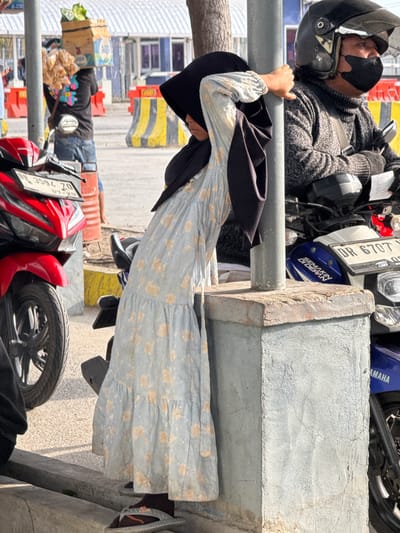 A young girl, wearing a light blue floral dress and a black hijab, leans against a concrete pillar near a street. She is positioned to the left of the pillar, with her right arm resting on it. A man wearing a helmet, mask, and a grey knitted sweater is visible to her right, seated on a blue and white motorcycle. A red motorcycle is parked to the far left of the frame. In the background, there are yellow and black striped barriers, suggesting a construction or traffic control area, and buildings that appear to be part of a commercial or transportation hub. The ground is paved with asphalt and a concrete curb is visible in the foreground. The scene suggests a public outdoor space, possibly in a marketplace or near a busy road. The location is identified as Upt Tambak Sari, Indonesia.