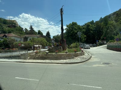 The image depicts a rural or small-town road intersection in Melnik, Bulgaria, under a clear blue sky with scattered white cumulus clouds.

In the foreground, an asphalt road with a white dashed lane marking leads to a central, irregularly shaped landscaped island. This island is bordered by stone curbing and contains grass, a large central rock formation, and several potted plants with red and white flowers. Rising from the center of the island is a tall, dark utility pole, from which numerous overhead power or communication lines extend horizontally across the upper portion of the frame. Attached to this pole are various conduits and pieces of electrical equipment. A gray sign with white text, reading "ЦЕНТЪР / CENTRUM", is visible near the utility pole.

To the left of the island, an asphalt road extends past a metal fence. Beyond this fence, residential buildings with white facades and red tile roofs are nestled among dense green foliage on a rising hillside. Higher on the hill, an unfinished or derelict brick structure with an exposed roof skeleton is visible. A red circular "No Entry" sign is positioned near the metal fence.

To the right of the island, an asphalt road curves uphill. Several parked vehicles are visible along this road, including a silver hatchback with a Bulgarian license plate "B 4051 PP". The right side of this road is bordered by a low stone wall painted with horizontal stripes of white, green, and red, representing the Bulgarian flag. Further along this road, a cluster of road signs is present: a red octagonal "STOP" sign, a blue circular "No Stopping/Standing" sign, a blue square "straight ahead" arrow sign, a blue square "turn right" arrow sign, and a red circular "20 km/h speed limit" sign. Below these signs, a "Hotel Melnik" sign with a phone number "+3598879131" is displayed. Beyond the road signs, some commercial stalls with light-colored awnings are discernible.

The background is dominated by densely forested hillsides with a mix of deciduous and coniferous trees. On the summit of the highest visible hill, a communication tower or antenna structure is present. No people are visible within the frame.