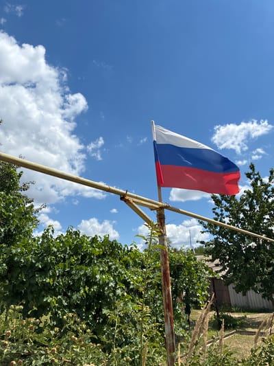 In Zhirnov, Russia, a Russian flag flies on a makeshift pole against a bright blue sky with scattered white clouds. The flag, consisting of white, blue, and red horizontal stripes, is attached to a rusty metal pole that is part of a simple, weathered structure. Behind the flag, lush green foliage, likely from trees and bushes, fills the lower half of the frame. In the background, a section of a building with a grey roof and walls is visible, suggesting a rural or suburban setting. The overall scene depicts an outdoor environment under a sunny day.