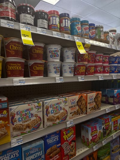 The image displays a detailed view of three merchandise shelves inside a retail establishment, likely a supermarket or pharmacy, located in Bal Harbour Village, United States. The top shelf features various jars of preserves and spreads, specifically Smucker's and Bonne Maman jams, Jif and Skippy peanut butter, and Nutella. The middle shelf is stocked with multiple rows of individual-serving cereal cups, including Honey Nut Cheerios, Cinnamon Toast Crunch, Lucky Charms, Froot Loops, and Frosted Flakes. The bottom shelf showcases an assortment of boxed snack bars and treats, with visible brands such as Cinnamon Toast Crunch Treats, Chex Mix Peanut Butter Chocolate, Rice Krispies Treats, Fruity Pebbles Treats, Quaker Chewy Yogurt, and Nature Valley Granola. Price tags are affixed to the shelf edges below several products. The environment is brightly lit, indicative of a typical retail interior. No individuals are present within the frame, and no actions or interactions are depicted.