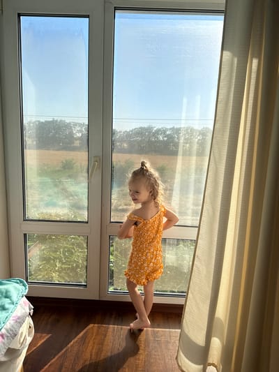 In a room with wooden flooring, a young girl with blonde hair tied in a messy bun stands facing a large window. She is wearing an orange, floral-patterned romper. Her back is to the viewer, and she is looking to the right with a smile. Sunlight streams through the window, casting shadows on the floor. Outside the window, a landscape with trees and fields is visible under a clear sky. To the right of the window, a beige curtain hangs, partially obscuring the view. In the lower left corner of the image, a laundry basket filled with blankets is visible. The scene is indoors, near a window that offers a view of an outdoor environment in or around Kosivka, Ukraine.