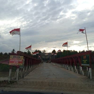 The image depicts a straight-on, low-angle view of the Teupin Reudeup bridge in Awegeutahpaya, Indonesia, from the perspective of a vehicle entering or traversing it. The bridge features a red-painted metal truss structure on both sides, supporting a roadway constructed from grey metal plates or concrete slabs, some showing signs of patching and wear. Numerous red and white flags, characteristic of the Indonesian national flag, are affixed to poles along both sides of the bridge, many appearing wind-blown and some visibly tattered.

On the left side of the bridge, a yellow and red sign with black text indicates "JEMBATAN TEUPIN REUDEUP," states a "BERAN MAKSIMAL 10 TON" (maximum load 10 tons), and mentions "YONZIPUR 16 DA" (Combat Engineer Battalion 16th Detachment/Area). Beneath the bridge on the left, a brown river is visible, flanked by dense green vegetation.

On the right side of the bridge, a green and yellow sign with white text also identifies the "JEMBATAN TEUPIN REUDEUP," mentions "ACEH BEREGEN," and specifies "10 DESEMBER 2016," followed by the slogan "NEGARA HADIR UNTUK RAKYAT" (The State is Present for the People).

In the distance, a white car is visible on the bridge, approaching the viewer. Beyond the bridge's structure, the surrounding environment consists of dense tropical foliage, primarily palm trees, extending under an overcast sky with scattered, bright clouds.
