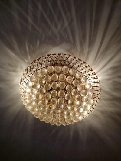 The image shows a close-up, low-angle shot of a chandelier hanging from a ceiling. The chandelier is round and appears to be made of brass or a similar metallic material, with numerous clear, faceted crystal spheres arranged in a spherical pattern. These spheres catch and refract the light, creating a shimmering effect and casting intricate patterns of light and shadow onto the ceiling above. The light source within the chandelier is visible, illuminating the crystals and casting bright highlights. The ceiling is dark, with the light from the chandelier creating dramatic, radiating beams and abstract shapes. The overall impression is one of elegance and luxury, with the play of light and shadow adding a dramatic and somewhat ethereal quality to the scene. There is no text visible in the image, and no indication of the time of day or weather conditions. The setting appears to be indoors, likely in a room with a high ceiling, given the way the light is projected. No people are visible.