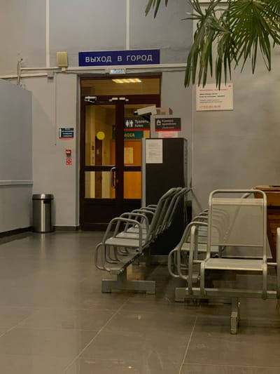 The image depicts an interior hallway, likely at a transportation hub, in Balezino, Russia. A blue sign overhead reads "ВЫХОД В ГОРОД" (Exit to the City), with a smaller green sign below it saying "ВЫХОД" (Exit). To the right of the exit doors, a white sign with red lettering states "Уважаемые пассажиры! Здесь всегда чисто! Если не согласны, звоните." (Dear passengers! It is always clean here! If you disagree, call.) Below this text is a phone number: "+7-910-405-46-00". Further to the right, another sign indicates "Туалет / Toilets" and "Камера хранения / Cloakroom". The hallway features several rows of empty, utilitarian metal chairs for seating. A silver trash receptacle stands near the left wall. The floor is tiled in a dark, neutral color. A potted palm tree with long fronds is visible in the upper right corner of the frame. The lighting appears to be artificial, casting a somewhat dim glow on the scene.