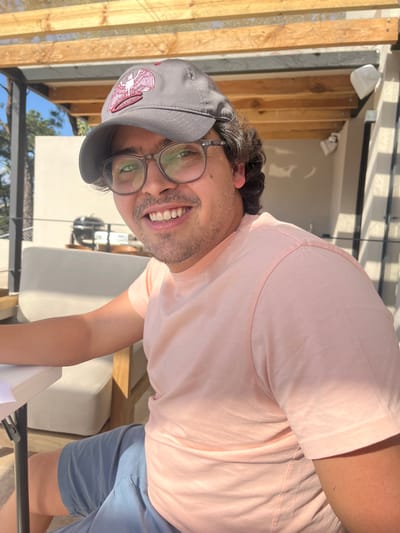 A man with dark curly hair smiles at the camera, wearing glasses and a gray baseball cap with a maroon design. He sports a light pink t-shirt and blue shorts. The background features wooden slats and a white exterior wall with visible fixtures. The man appears to be sitting or standing outdoors on a sunny day.