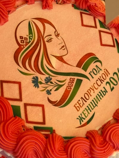 This is a close-up, top-down view of a cake, likely decorated for a celebration. The cake has an edible image on its surface depicting a stylized profile of a woman with long, flowing hair in shades of red, green, and brown. Accompanying the portrait is text in Russian: "ГОД БЕЛОРУССКОЙ ЖЕНЩИНЫ 2020", which translates to "Year of Belarusian Women 2020". There are also abstract geometric patterns in red and green squares on the cake's surface, and a small cluster of blue flowers. The edges of the cake are adorned with red frosting piped in a ruffled or wave-like pattern. The image does not provide information about its location in Вяйнянскі сельскі Савет, Belarus.