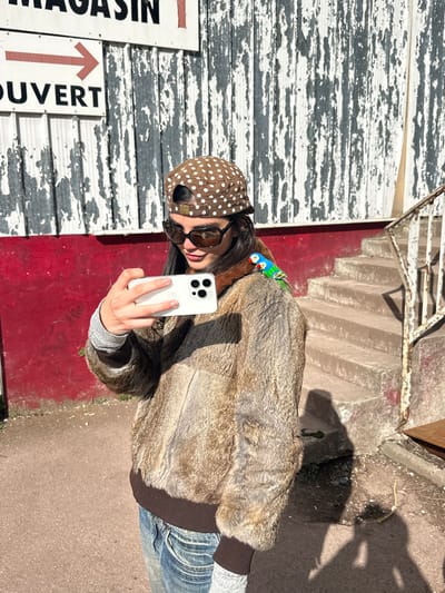 An individual, appearing to be a young woman, stands outdoors in Neuilly-Plaisance, France, captured in a sunlit, mid-shot frame. She wears a brown baseball cap with white polka dots, reversed with the brim facing backward, and large dark sunglasses. Her attire includes a light brown faux fur bomber jacket with dark brown ribbed cuffs and hem, a grey long-sleeved shirt underneath, and blue jeans. Her right hand holds a white smartphone with a prominent triple-lens camera array, positioned to capture a self-portrait or video, indicated by its orientation towards herself. A small, vibrantly colored bird, predominantly blue, green, and red, is perched on her right shoulder.
The background features a building with a distinct two-toned facade. The lower section is painted a solid dark red. Above this, the wall consists of distressed, peeling white paint over a darker surface, possibly wood or metal siding, revealing a worn texture. Two white rectangular signs are affixed to this upper section; the top sign partially displays "MAGASIN" (store), while the lower sign features a red arrow pointing right and the word "OUVERT" (open). To the right of the individual, a set of weathered concrete or stone stairs ascends, accompanied by a simple, light-colored metal handrail. The ground beneath the subject is a reddish-brown paved surface, possibly asphalt. Strong natural light casts shadows on the ground to the right.