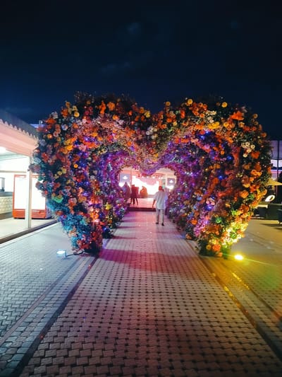 The image captures an enchanting nighttime scene in Aksukent, Kazakhstan, featuring a massive heart-shaped archway meticulously adorned with an abundance of colorful flowers and twinkling lights. The archway creates a tunnel-like effect, inviting viewers to walk through it. A pathway paved with interlocking bricks leads towards the archway, with subtle lighting illuminating the ground.
At least three people are visible, one walking away from the viewer and into the heart-shaped structure, dressed in a white shirt and light-colored trousers. Another figure, also dressed in light-colored clothing, is seen inside the archway. The scene is set outdoors, under a dark, cloud-speckled night sky. The lighting is primarily artificial, with vibrant blues, purples, oranges, and yellows emanating from the decorative lights entwined with the flowers, casting a warm glow on the surroundings.
On the left side of the image, a building with a white facade and a series of windows or entrances is partially visible. Red vending machines are positioned near one of the entrances, suggesting a commercial or public space. The overall atmosphere is festive and romantic, evoking a sense of wonder and celebration. The time of day is clearly night, indicated by the dark sky and artificial lighting. No visible text is present in the image.