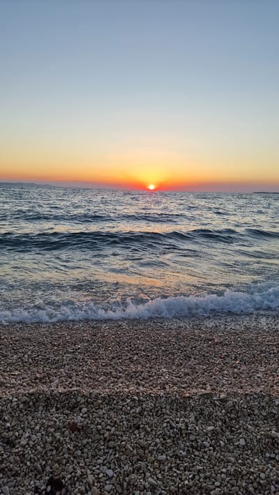 The image depicts a serene beach scene at sunset, with the sun low on the horizon casting a warm, golden glow. The sky transitions from a soft blue to a vibrant orange as it nears the sun. The sea is calm, with gentle waves lapping at the shore. The beach itself is covered in small, smooth pebbles, creating a textured foreground. The overall atmosphere is peaceful and tranquil, evoking a sense of calm and relaxation. The time of day is clearly sunset, and the lighting suggests fair weather. There are no people or man-made objects visible in the frame, focusing solely on the natural beauty of the coastal landscape. The location is Zadar, Croatia.