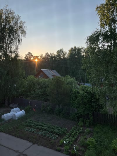 A wide shot captures a backyard scene in Yaransk, Russia, during sunset. The sky is a pale blue, transitioning to a warm orange glow as the sun dips below a line of trees in the distance. Sunlight pierces through the trees, creating visible rays. A small, wooden house with a grey roof is visible amidst the foliage. A garden occupies the foreground, with rows of green plants, including what appear to be cabbage and strawberries. Two white hoop-shaped greenhouses cover some of the garden area. A dark wooden fence runs horizontally across the middle ground, separating the garden from the trees and the house. The overall environment appears rural and tranquil.