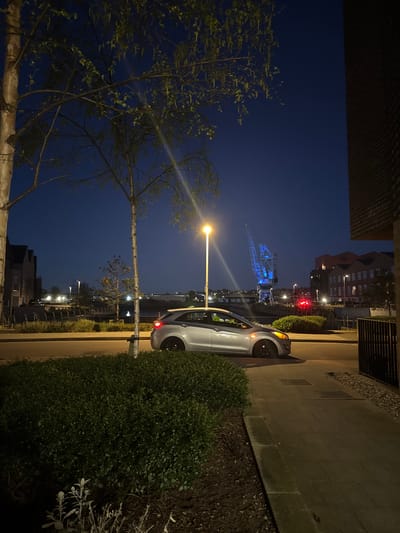 The image depicts a nighttime scene in Rochester, United Kingdom. A silver car is parked on a paved street next to a building and a substantial hedge. In the distance, a brightly lit blue crane, a local landmark, is visible against the dark sky. Streetlights illuminate the scene, casting light on the buildings and foliage. A tree with sparse leaves is on the left side of the image. The sky is a deep blue, suggesting it is late evening or night. The location appears to be a residential area within the city, close to the riverfront.