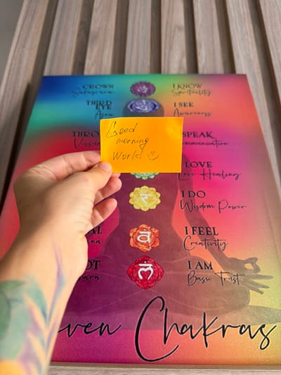 A hand is holding an orange sticky note with the words "Good morning World :)" written on it in black marker. The sticky note is positioned over a large canvas print of the seven chakras. The canvas displays the chakras as colored circles with Sanskrit symbols, and lists associated affirmations such as "I KNOW Spirituality," "I SEE Awareness," "I SPEAK Communication," "I LOVE Love Healing," "I DO Wisdom Power," "I FEEL Creativity," and "I AM Basic Trust." The background of the canvas features a gradient of colors, from blue and green at the top to pink and yellow at the bottom, and a silhouette of a person in a meditative pose. The canvas is resting on a wooden surface, possibly a table or a floor, with vertical wooden slats visible in the background. The image does not provide any information about the location in Nha Trang, Vietnam.