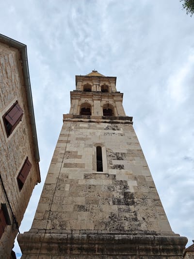 This is a low-angle, eye-level, wide shot of a stone bell tower in Grad Stari Grad, Croatia, against a cloudy sky. The tower, constructed of light-colored, weathered stone blocks, dominates the right side of the frame. It has multiple levels with arched openings, some of which are covered with dark grating. A thin black cable runs vertically down the side of the tower. To the left of the tower, a portion of a stone building is visible, featuring two windows with dark wooden shutters, one partially open. The building is also made of rough-hewn stone. The sky is overcast with visible cloud formations. There are no people or animals visible in the image.