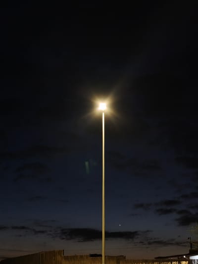 The image displays a nighttime scene in Montuïri, Spain, dominated by a single, centrally positioned street lamp. The lamp features a tall, slender, light-colored pole, possibly beige or pale yellow, topped with a rectangular light fixture that emits a bright, warm white illumination with a distinct starburst effect. The sky above is dark, transitioning from deep black in the upper frame to a dark blue-grey closer to the horizon, with scattered dark, indistinct clouds. A small, bright white point of light, consistent with a star or planet, is visible in the sky to the right of the lamp pole. A faint, greenish vertical light streak or artifact is present in the upper-left quadrant of the frame, below the lamp's light source. At the bottom of the image, horizontally oriented structures are visible: a textured, possibly concrete or masonry wall extends across the lower-left, and to the right, a darker, less defined structure with a hint of sparse foliage. No individuals, animals, or active events are depicted.