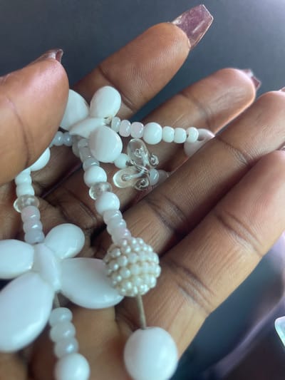 The image shows a close-up view of a person's hand holding a white beaded necklace. The hand has dark skin and is adorned with long, clear acrylic nails. The necklace is made up of various white beads, including small seed beads, larger round beads, and flower-shaped charms. There are also a few clear, butterfly-shaped charms interspersed among the beads. The background is dark and out of focus. The lighting suggests an indoor setting, possibly with artificial light. There is no visible text in the image.