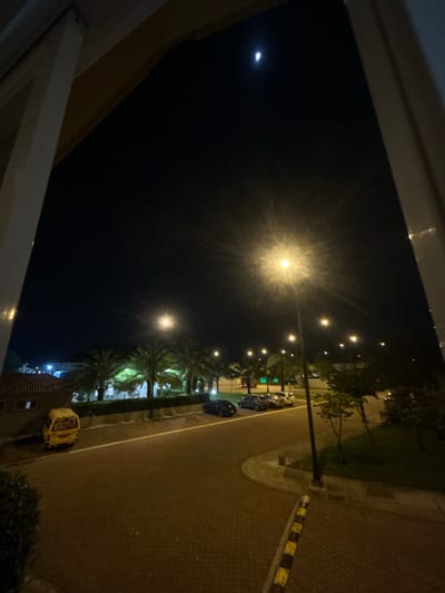 The image shows a night scene taken from an elevated position, likely from inside a building looking out a window. The sky is dark with a visible moon in the upper left. Two bright streetlights are visible. Palm trees line the street. Cars are parked along the street. The road is paved with bricks. There are buildings near the bottom. Based on the prompt, this is in La Aurora, Ecuador.