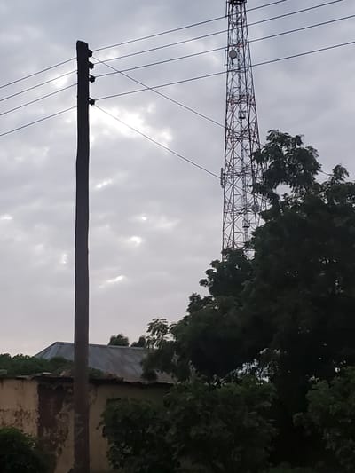 The image depicts a view of Kabwir, Nigeria, on a cloudy day, featuring a tall, lattice-style communication tower. In the foreground, a wooden utility pole with multiple wires stretches upwards. To the left, a portion of a building with a corrugated metal roof and a weathered facade is visible. Dense green foliage obscures much of the lower portion of the scene, with trees extending upwards to partially surround the communication tower. The sky is overcast with visible cloud formations.
