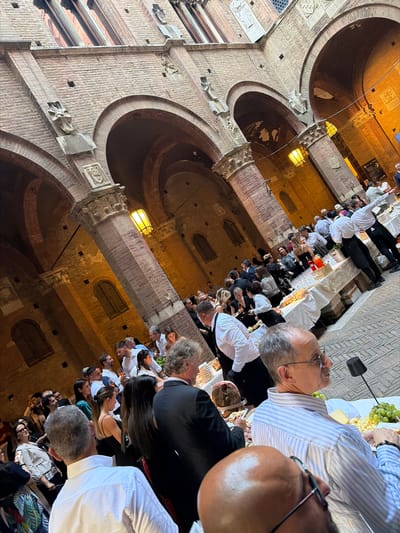 The image depicts a crowded event held in a historic courtyard, likely in Siena, Italy, given the distinctive brick architecture with arches and ornate column capitals, reminiscent of the Palazzo Pubblico or similar medieval structures. A long buffet table laden with food, including cheeses and cured meats, is set up, with servers in white shirts and black aprons attending to guests. People are mingling, with some appearing to be eating or drinking. The lighting suggests it is evening, with warm yellow lights casting a glow on the brick walls and illuminating the scene. The overall atmosphere suggests a festive gathering or reception.