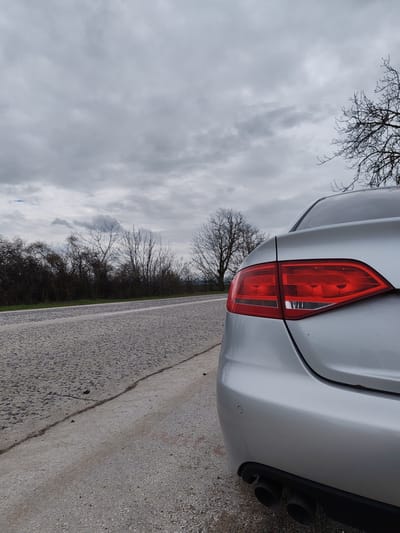 The image shows the rear of a silver sedan parked on the shoulder of a paved road. The car's right taillight is prominently featured. The road is cracked and worn, with a line indicating the edge of the paved surface. Beyond the road is a low-lying area with bare trees and bushes. The sky is overcast with heavy clouds. The location is Borovan, Bulgaria.