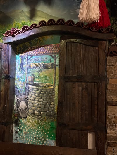 The image depicts a mounted display, possibly within a restaurant. At the top, there is a cabinet made of dark-stained wood with two hinged doors secured by a central clasp. Above the cabinet, a wall made of roughly hewn stones is visible. Below the cabinet is a painting, framed by a dark wood casing with decorative scallops. The painting depicts a scene with a well and a donkey; the perspective is as if the viewer is looking down into the well. Adjacent to the frame, on the upper left, is a decorative fringe of white and red yarn. The overall aesthetic suggests a rustic, European-themed establishment.
Given the specific request for a location in Des Plaines, United States of America, and knowing the decor style, it may be located in a restaurant or tavern there.