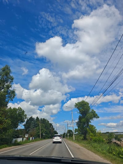The image depicts a view from a vehicle traveling on a paved road through a rural area, likely the village of Repnikovo, Russia, on a sunny day with scattered clouds. The road has white lane markings and a shoulder. A white hatchback car is visible ahead on the road, traveling in the same direction. The landscape is characterized by lush green trees and vegetation on either side of the road. In the foreground, the dashboard of the vehicle from which the photo is taken is visible. A power line runs along the right side of the road, supported by utility poles. A circular sign with Cyrillic text is affixed to a fence on the left side of the road. The sky is a vibrant blue, with numerous white cumulus clouds creating a dynamic and scenic backdrop.