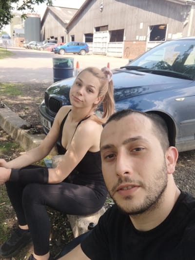 The image shows a selfie of a man and a woman. The man has short dark hair, a beard, and is wearing a black t-shirt. The woman has blonde hair pulled back in a ponytail and is wearing a black top and leggings. They are positioned near a blue car, partially visible in the frame. The car appears to be parked in an area outside a barn-like structure with a series of rectangular windows. Other vehicles, including a blue one, are visible in the background. The photo was likely taken in Stafford, United Kingdom, given the location.