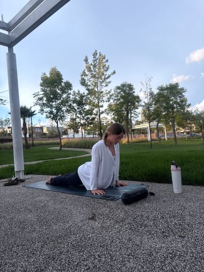 A young woman with braided brown hair is performing a yoga pose on a mat outdoors. She is wearing a white long-sleeved shirt and dark leggings, with her body in a Cobra pose, torso raised and hands on the mat for support. Her face is angled downwards, looking towards her hands. Beside her on the mat are a smartphone and a black cylindrical object, possibly a portable speaker or yoga block. To the right of the mat, a tall white water bottle with a pink lid stands upright. The woman is positioned on a textured gray ground composed of small pebbles. In the background, a lush green lawn is visible, bordered by various trees and modern buildings with clean architectural lines, suggesting an urban or suburban park setting. A large grey metallic structure, likely part of a park amenity or canopy, extends from the left side of the frame over the scene. The sky above is a clear light blue with a few wispy white clouds. The overall environment appears to be the city of Tlaxcalancingo, Mexico, likely during daylight hours with soft natural lighting.