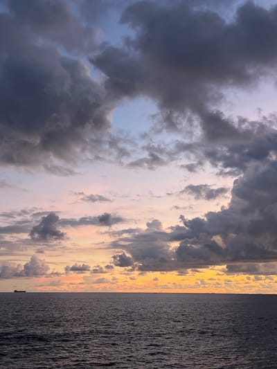 The image displays a dramatic and colorful sunset or sunrise over the ocean, likely taken from a vessel or a coastal vantage point in Rumassala, Sri Lanka. The sky occupies the upper portion of the frame, dominated by a dynamic interplay of clouds. Heavy, dark grey and purplish clouds fill much of the scene, appearing dense and voluminous. Between these darker masses, lighter clouds catch the warm glow of the low sun, exhibiting bright golden, orange, and soft pink hues, especially closer to the horizon. Patches of pale blue and lilac sky are visible through breaks in the cloud cover.

The lower half of the image is filled by the vast, dark expanse of the ocean. Its surface is textured with countless ripples and small waves, creating a consistent pattern. A clear horizon line separates the water from the sky. On this distant horizon, a very small, dark silhouette of what appears to be a boat or ship is faintly discernible. The overall atmosphere is one of serene power and natural beauty, characteristic of dawn or dusk at sea. No visible text is present in the image.